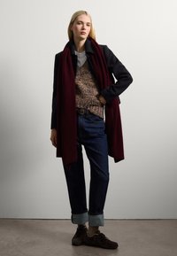 Une écharpe bordeaux drape un pull en maille brun texturé, associé à un jean bleu foncé et des chaussures marron. L'arrière-plan est gris clair.