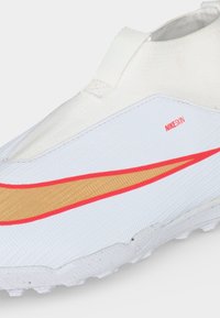 Λευκό αθλητικό παπούτσι Nike slip-on με χρυσό σήμα swoosh περιγεγραμμένο με κόκκινο, ανάγλυφο επάνω μέρος και μικρή κόκκινη ετικέτα NIKESKIN στο πλάι.