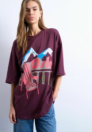 Giovane donna che indossa una maglietta viola oversize con una grafica di montagne, sedia e la scritta "Oslo", abbinata a jeans blu, in piedi davanti a uno sfondo bianco.