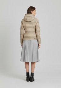 Beige donzen jas met horizontaal doorgestikt patroon, capuchon en logodetail, gedragen over een geplooide grijze rok en zwarte enkellaarsjes met hak.