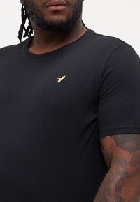 Schwarzes T-Shirt mit kurzen Ärmeln aus weichem Stoff, das auf der linken Brustseite ein kleines, goldgesticktes Logo aufweist.
