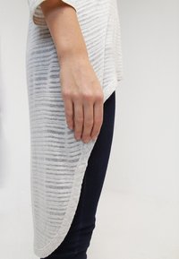 Krémový a šedý proužkovaný pletený top s zakřiveným lemem, zkombinovaný s tmavě modrými džínami. Tkanina působí měkce a lehce, ukazující umístění ruky.