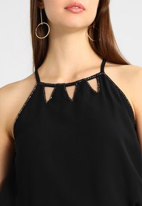 Haut noir sans manches avec col halter, présentant des découpes triangulaires et des accents métalliques cloutés le long de l'encolure et des emmanchures.
