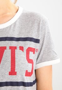 Camiseta de algodón gris con mangas cortas, ribete blanco y un logo rojo de "LEVI'S". Presenta rayas horizontales en azul marino debajo del logo.