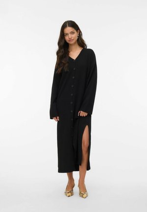 Robe cardigan longue noire avec un décolleté en V et une fermeture à boutons, dotée de manches longues et d'une fente sur le côté, portée avec des chaussures à bout pointu à motifs.