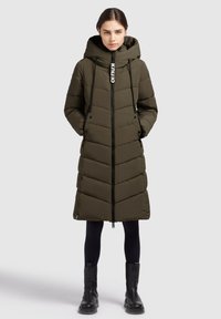 Garšs olīvzaļš pūkains mētelis ar čevronu šuvēm, augstu apkakli, auklēm, priekšējo rāvējslēdzi un sānu kabatām. Valkāts virs melnajām legingām un zābakiem.