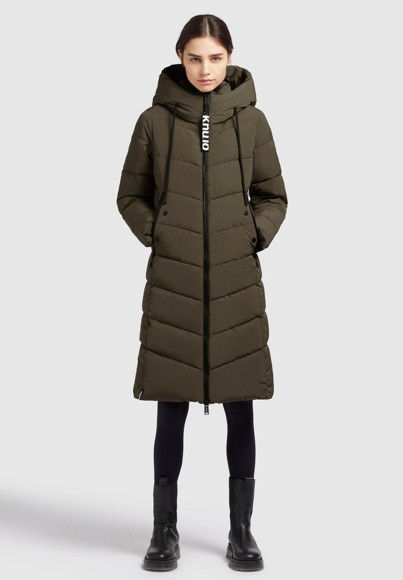 Garšs olīvzaļš pūkains mētelis ar čevronu šuvēm, augstu apkakli, auklēm, priekšējo rāvējslēdzi un sānu kabatām. Valkāts virs melnajām legingām un zābakiem.