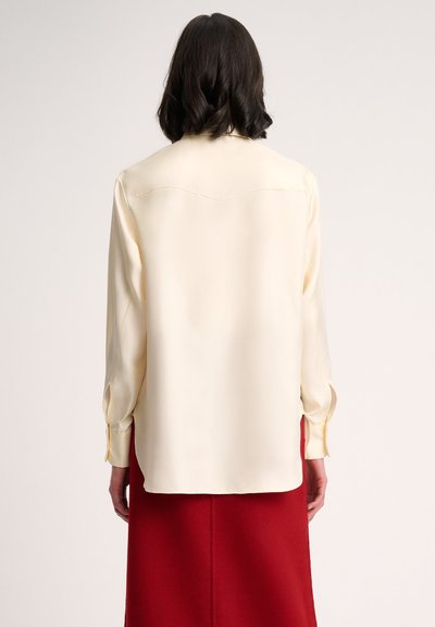 Blusa di satin color crema chiaro con maniche lunghe, orlo arrotondato e sottile cucitura sul retro. Indossata con una gonna rossa, che mette in evidenza la sua consistenza liscia.
