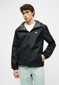 Νεαρός άνδρας με σγουρά μαλλιά που φοράει μαύρο φούτερ με φερμουάρ και ανοιχτ όχρωμο παντελόνι, στέκεται με το ένα χέρι στην τσέπη.