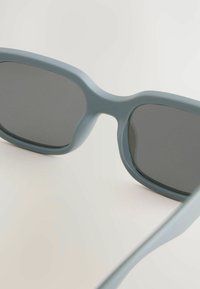 Lunettes de soleil teintées de gris avec une monture ronde en plastique mat bleu clair, dotées de branches légèrement courbées et d'une texture lisse.