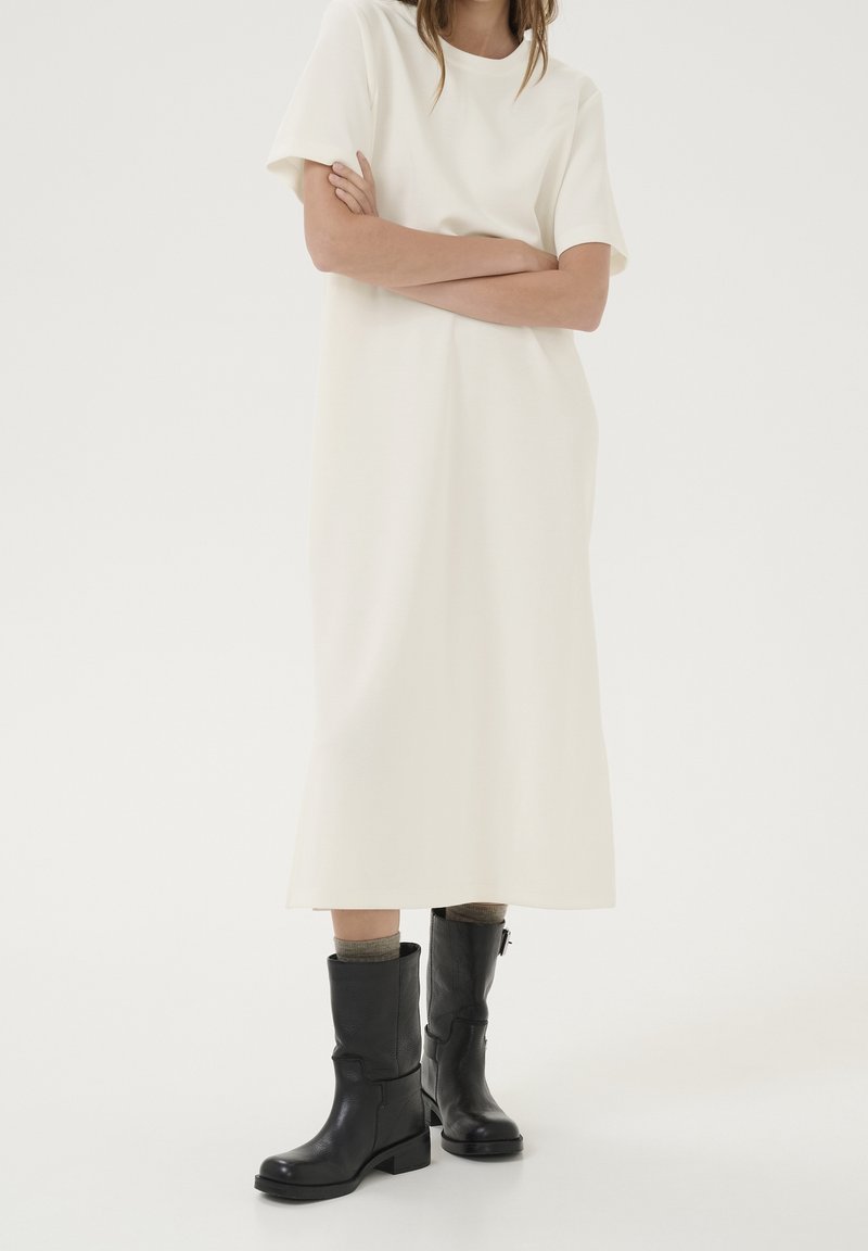 Femme debout, les bras croisés, portant une robe midi blanche et des bottes en cuir noires mi-mollet avec des chaussettes grises sur fond blanc.
