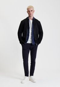 Giacca bomber in suede nera indossata sopra una camicia di denim, abbinata a un maglione bianco, pantaloni blu navy e sneakers bianche.