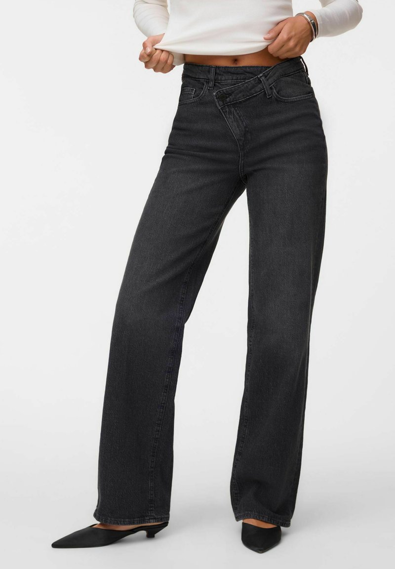 Femme portant un jean noir taille haute à jambes larges avec une fermeture asymétrique à boutons, associé à des talons noirs pointus et un haut blanc.