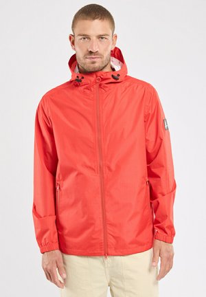 Homme portant une veste coupe-vent à capuche rouge vif avec fermeture éclair frontale et pantalon beige sur fond blanc uni.