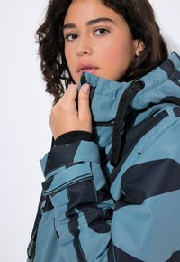Giacca impermeabile in teal e nero, con colletto alto, zip e polsini elasticizzati. Texture liscia con motivi geometrici.