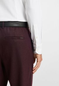 Witte, geribbelde lange mouw shirt gecombineerd met donkere bordeaux, gestructureerde broek. Voorzien van een zwarte leren riem en discrete branding op de zak.