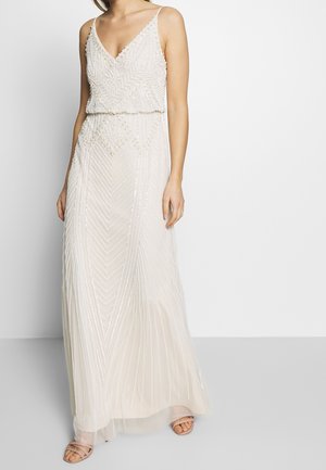 Kobieta ubrana w bezrękawnikową białą sukienkę z ozdobami z koralików i cieliste sandały na paski, stojąca na tle o jednolitym kolorze.