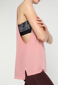 Roze, mouwloze atletische tanktop met een geperforeerde textuur, zijsplitten en een losse pasvorm, gelaagd over een zwart patroon sportbeha.