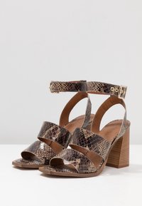 Högklackade sandaler med ett orm-mönster, med två korsande remmar, ankelrem och en träblockklack. Färgerna är bruna och beige.