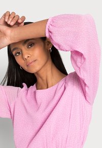 Blusa rosa com mangas bufantes, apresentando um tecido texturizado e pequenos pois brancos, gola redonda e detalhes franzidos nos ombros.