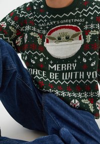 Pull en maille verte avec un motif graphique d'un personnage portant un chapeau de Père Noël, entouré de flocons de neige et de texte festif en blanc et rouge.