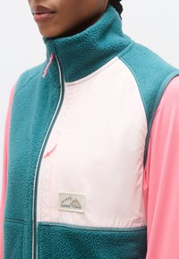 Γιλέκο από φλις σε τιρκουάζ και ανοιχτό ροζ, με ψηλό γιακά, πάνελ από αντίθετο ύφασμα και ένα μικρό μπατζάκι με σχέδιο βουνού.