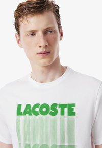 Joven con el pelo corto y rizado de color rojo, vistiendo una camiseta blanca con el texto distorsionado "LACOSTE" en verde sobre un fondo blanco sencillo.