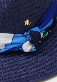 Καπέλο από άχυρο σε ναυτικό μπλε χρώμα με υφασμάτινη ταινία σε μπλε και λευκό σχέδιο, διακοσμημένο με χρυσό λογότυπο "RL". Υφαντή υφή και λεπτομερής σχεδιασμός.
