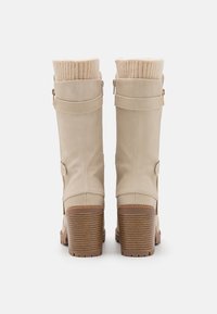 Botas de tobillo de cuero sintético beige con puños de punto acanalado, cremalleras laterales y tacones apilados que presentan bases de color marrón texturizado y accesorios de hardware dorado.