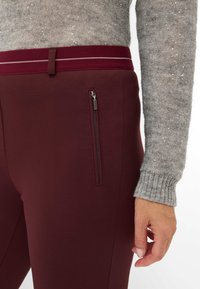 Burgunderfarbene Strickhosen mit einer figurbetonten Passform, ausgestattet mit einer seitlichen Reißverschluss-Tasche und einem kontrastierenden weinroten Bund mit weißen Details.