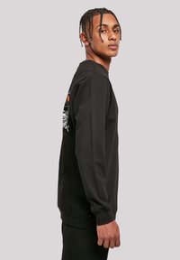 Junger Mann mit geflochtenem Haar, der ein schwarzes Langarmshirt mit grafischem Design auf dem Rücken trägt, im Profil vor einem schlichten Hintergrund stehend.