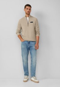Beige Zip-Kragen-Pullover mit strukturiertem Strick, hellblaue Jeans und crèmefarbene Sneaker mit braunen Akzenten. Das Model steht vor einem neutralen Hintergrund.