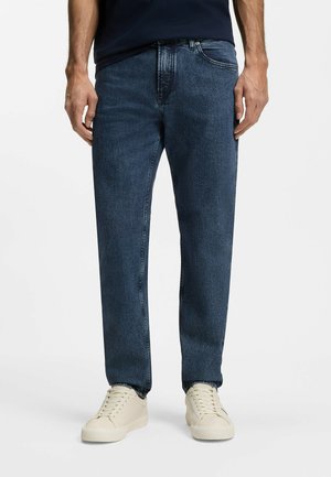 Mężczyzna noszący ciemnoniebieskie jeansy o prostych nogawkach i kremowo-białe sneakersy sznurowane, stojący na jednolitym białym tle.