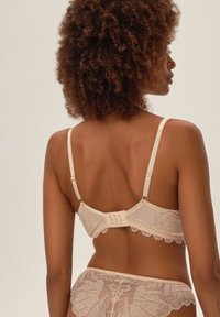 Beżowy biustonosz z koronką, z falbaniastym dołem i regulowanymi ramiączkami. Zapięcie z wieloma haczykami do regulacji dopasowania. Teksturowany wzór kwiatowy.