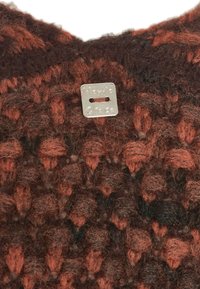 Maglione lavorato a maglia in colori marrone scuro e ruggine, caratterizzato da un motivo testurizzato e un'etichetta rettangolare argentata con un foro per il fissaggio.