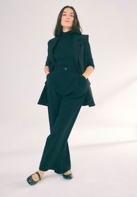 Svart skräddarsydd kavaj med hög krage över en åtsittande polokrage. Höga, vida svarta byxor. Prickiga slip-on skor.