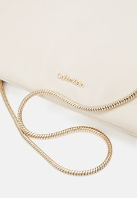 Beige dámská kabelka Calvin Klein z umělé kůže se zlatým řetízkem. Má hladký povrch a jemně embosované logo na přední straně.