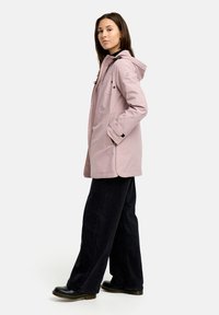 Hellrosa wasserdichte Jacke mit Kapuze, seitlichen Taschen, schwarzen Akzenten und lockerer Passform, kombiniert mit schwarzen weit geschnittenen Hosen und robusten Schuhen.