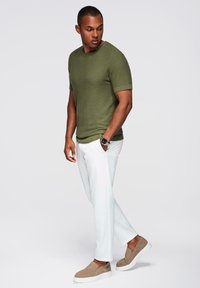 Olivově zelená texturovaná košile s krátkým rukávem, bílé úzké kalhoty a světle hnědé slip-on boty. Jednoduchý design s minimem kovových detailů.