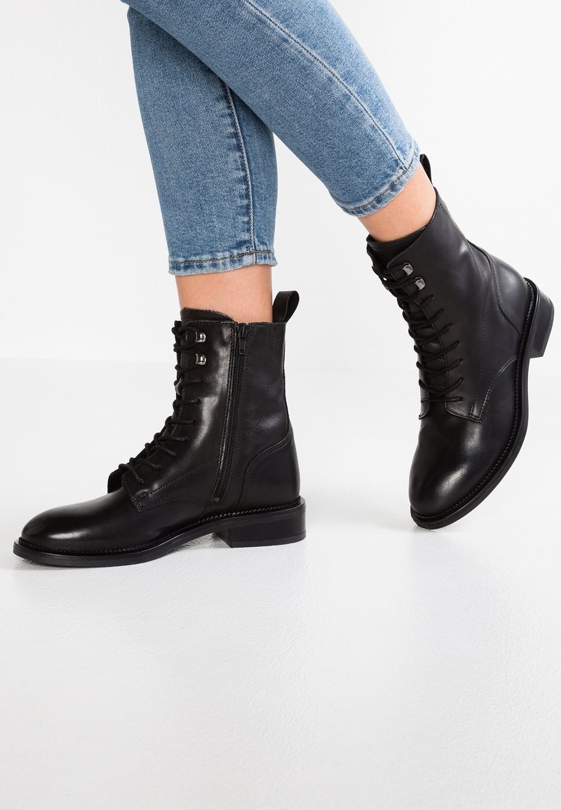 Jambes en jean bleu clair portant des bottes en cuir noir montant jusqu'au milieu du mollet, à lacets et zippées, un pied à plat, l'autre levé sur un sol blanc.