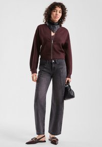 Vrouw in een bordeauxrode ritstrui, zwarte leren halssjaal, grijze rechte jeans, wijde pumps met studs, die een kleine zwarte handtas vasthoudt.