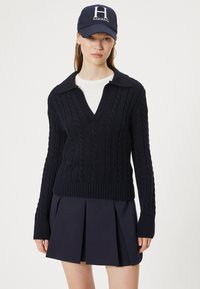 Marineblauwe gebreide trui met een V-hals en kabelpatroon, gecombineerd met een marineblauwe plooirok en een marineblauwe pet met een wit "Harvard" logo.