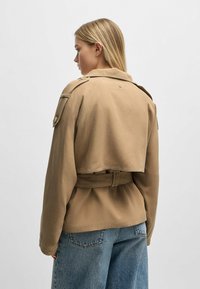 Tan short jacket with a belted waist, fold-over collar, and flap pockets. Soft fabric with a relaxed fit and subtle stitching details.