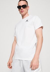 Fehér pamut póló, a bal mellen fekete Jumpman logóval; világosszürke sorttal és fekete napszemüveggel párosítva.