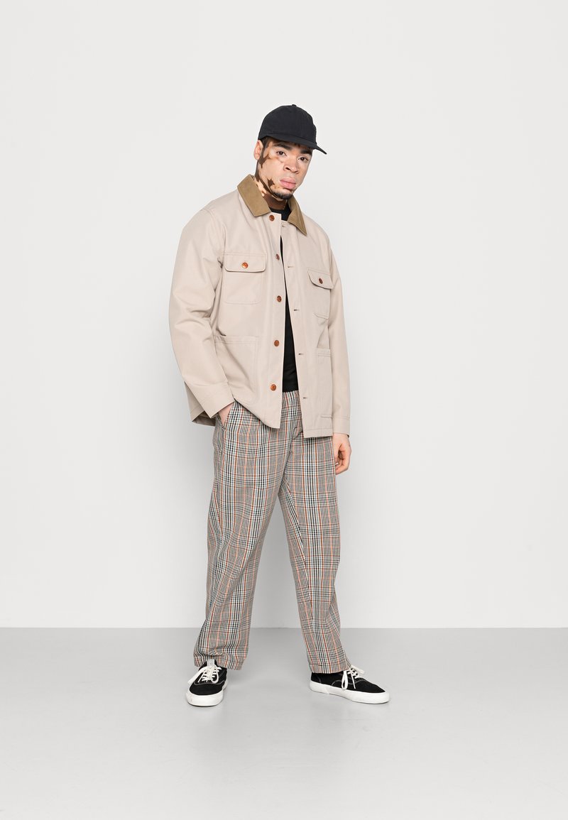 Giacca di cotone beige con colletto marrone, due tasche sul petto, chiusura a bottoni, abbinata a pantaloni a quadri bianchi, verdi e rossi, e sneakers nere.