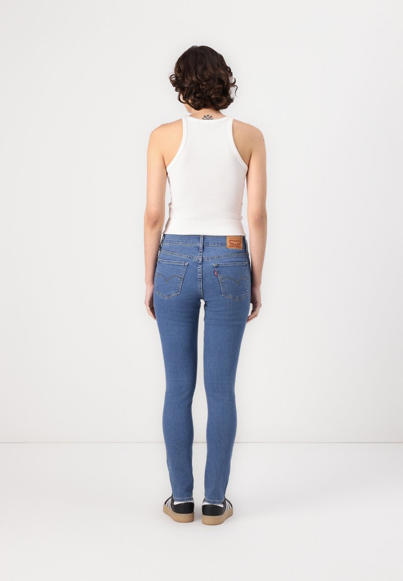 Femme aux cheveux bouclés portant un débardeur blanc, un jean skinny bleu et des baskets noires, se tenant devant un mur blanc uni.