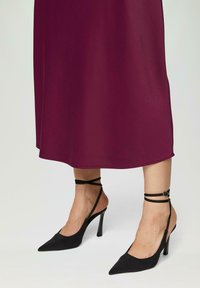 Zwarte high-heeled schoenen met een puntige neus en enkelbandjes, met een gladde textuur en een slank ontwerp, gecombineerd met een bordeauxrode jurk.