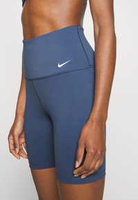 Zbliżenie osoby noszącej niebieskie sportowe spodenki Nike z wysokim stanem, z białym logo Nike Swoosh po lewej stronie.