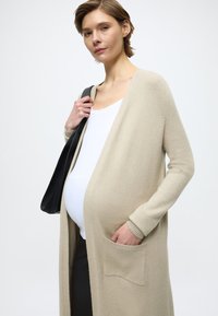 Beżowy sweter na drutach z długimi rękawami i otwartym przodem, z przednią kieszenią. Nosi się go na białym topie, z czarną torbą na jednym ramieniu.