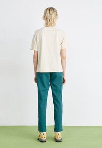 Beige kortermet t-skjorte, teal bukser og gule sneakers med svarte detaljer. Glatt stoff, avslappet passform, synlig søm på nederkant.